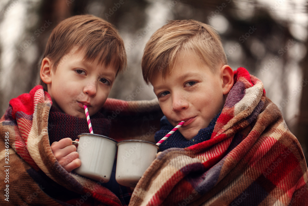 Two children bundled together in a plaid blanket outdoors, sipping mugs of Sillycow hot chocolate through striped straws