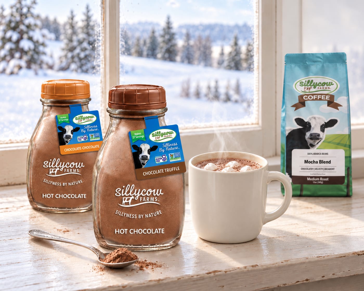 Sillycow Chocolate Truffle and Chocolate-Chocolate hot chocolate bottles on a farmhouse window sill beside a steaming mug of hot chocolate with marshmallows, a spoonful of cocoa and a bag of Sillycow Mocha Blend coffee, with snowy fir trees outside