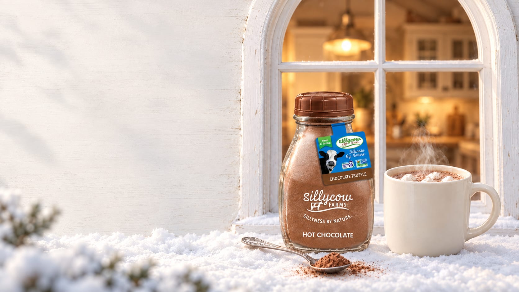 A Sillycow Chocolate Truffle hot chocolate bottle and a steaming mug of hot chocolate with marshmallows on a snowy window ledge, a spoonful of cocoa in front, with a warm lamplit farmhouse kitchen glowing through the arched window